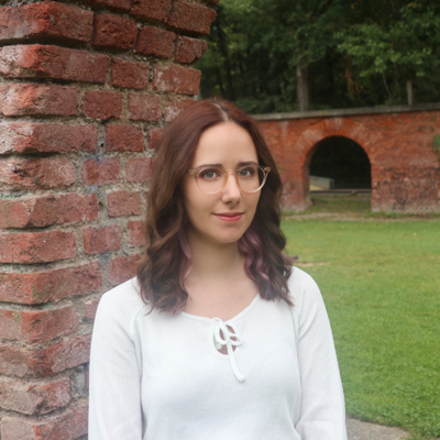 Foto von Christina Lari: Sie trägt eine Brille und steht vor einer Ziegelmauer