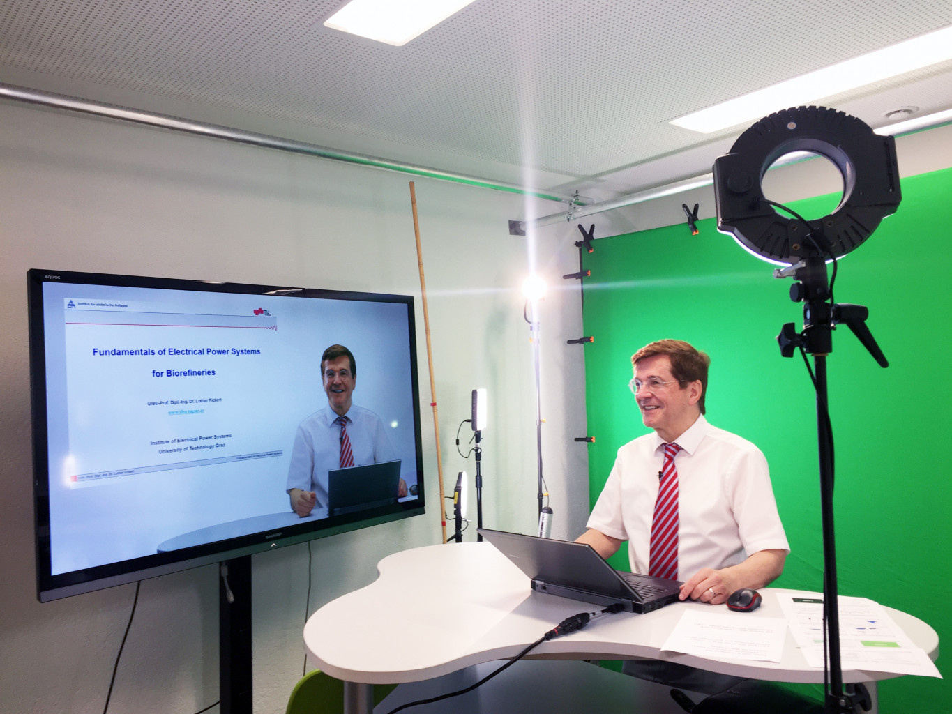 Videodreh im Studio. Ein Lehrender steht an einem Pult und sieht daneben einen Bildschirm mit einer Vorschau der Folien