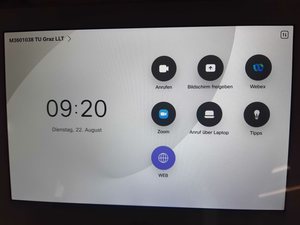 Foto des Touchpanels. Man sieht den Namen der Anlage, die Uhrzeit und die Menüpunkte Anrufen, Bildschrim Freigeben, Webex, Zoom, Anruf über Laptop, Tipps und Web