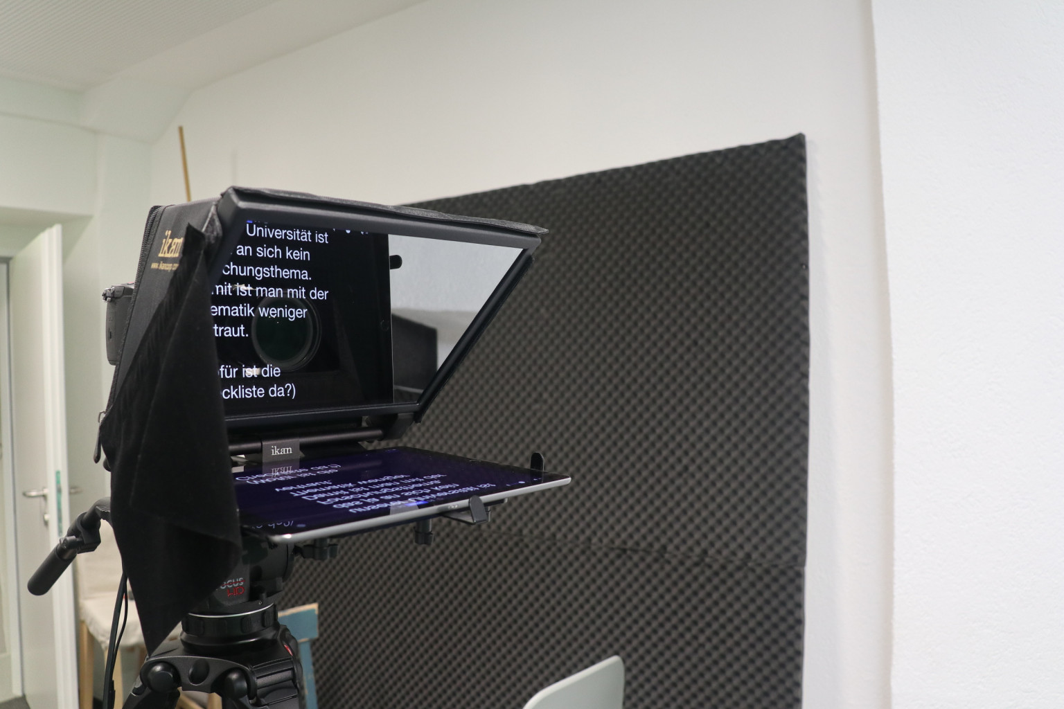 Teleprompter auf einem Stativ im Studio, auf der Spiegelscheibe läuft der Sprechtext, dahinter eine Akustikplatte an der Wand