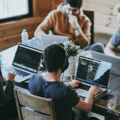 Mehrere Personen arbeiten mit Laptops an einem großen Holztisch, eine davon mit Kopfhörern