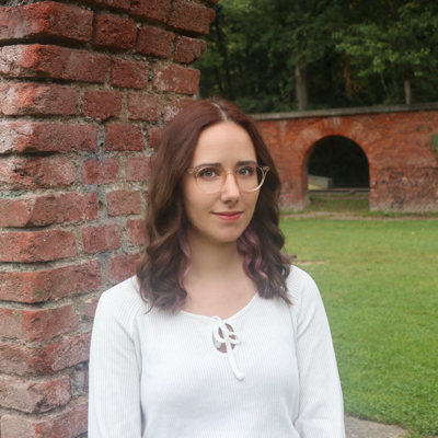 Photo of Christina Lari: She is wearing glasses and standing in front of a brick wall